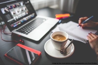 photo showing someone's hand writing in front of a notebook with a coffee on the desk.