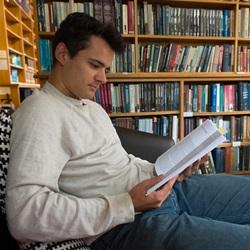 A seated person reading a book in a library setting with bookshelves behind