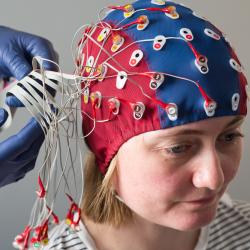 A person wearing an EEG cap on their head whilst a person behind attaches wires to the cap