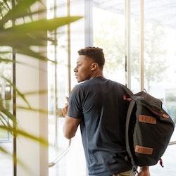 A black man looking outside window carrying a black and brown backpack.